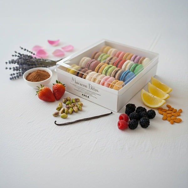 A 24-piece box of assorted French macarons surrounded by fresh ingredients including strawberries, raspberries, blackberries, lemon wedges, pistachios, vanilla bean, lavender, and cocoa powder.
